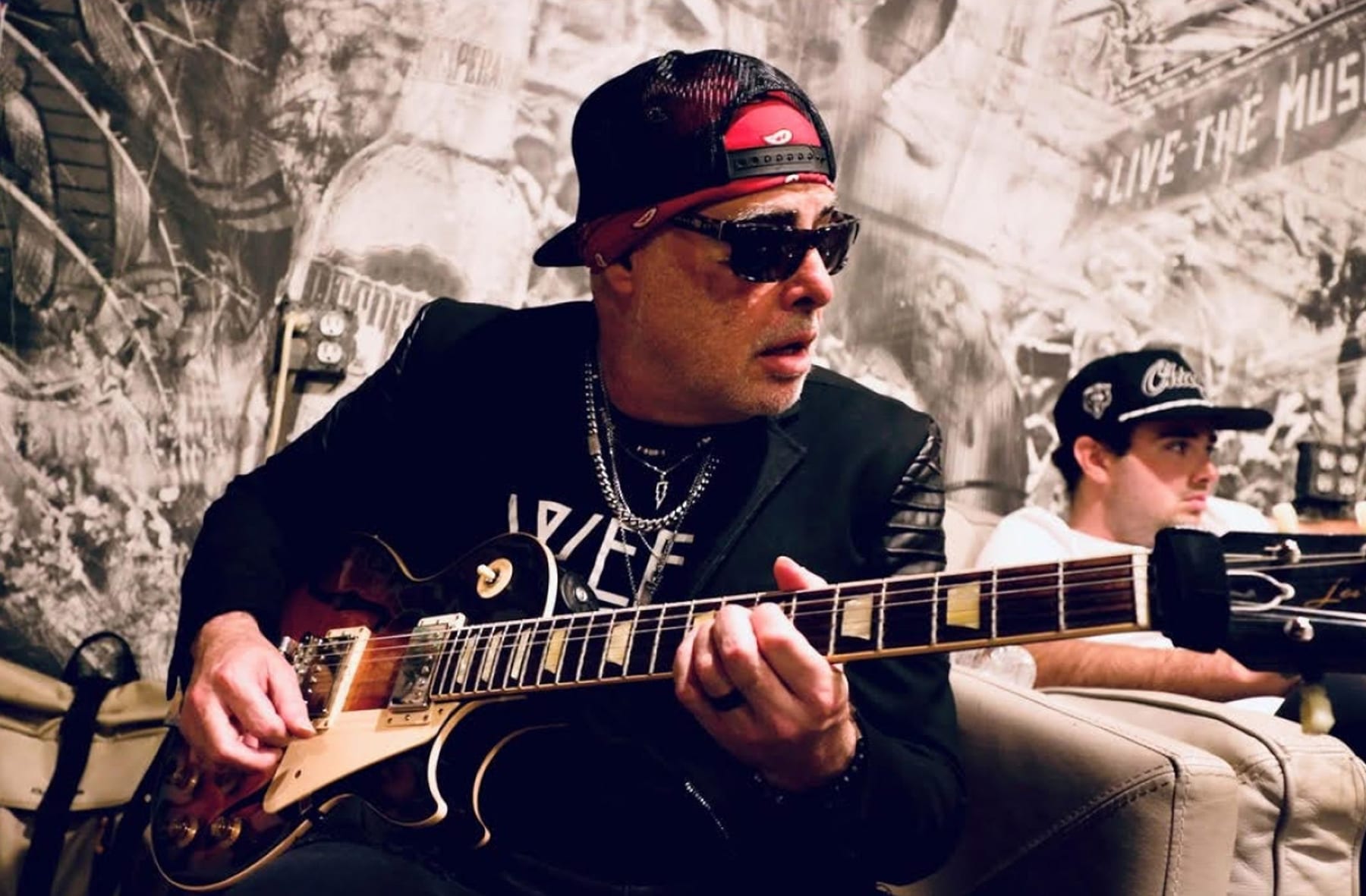 Alberto González playing a sunburst Les Paul backstage, wearing sunglasses, a backward cap and a bandana.