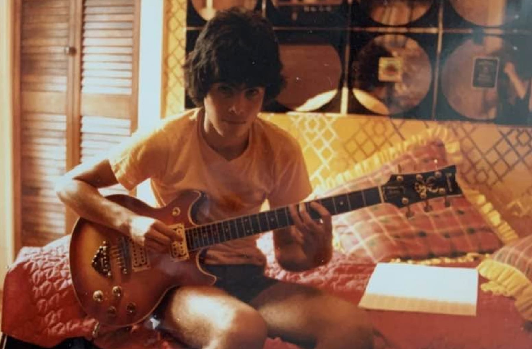 Young Alberto González seated on a bed playing a sunburst electric guitar in his bedroom, records on the wall behind him.